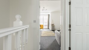 Un couloir avec une porte blanche, un escalier et une vue sur une pièce où se trouvent une chaise jaune et une lampe.