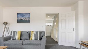 Un salon avec un canapé gris, un tableau représentant un port de plaisance, une lampe et un escalier menant à une autre pièce.