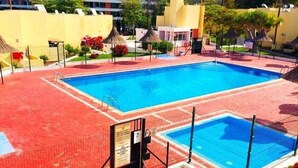 A swimming pool with a red tiled deck, surrounded by lounge chairs and umbrellas, with a building in the background. - El cortijo, pool view (Costa Adeje)