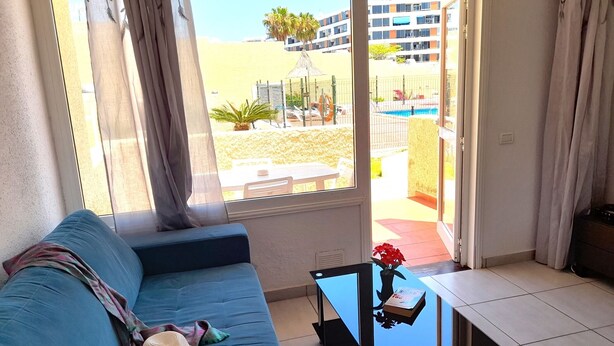A living room with a blue sofa, a small table with a vase, and a view of a pool and buildings outside. - El cortijo, pool view (Costa Adeje)