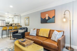 Een woonkamer met een bruine leren bank, een geel poufje, een houten salontafel en een schilderij aan de muur.
