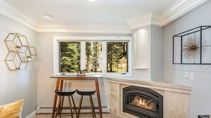 Una cocina moderna con chimenea, un área de comedor con dos taburetes y un amplio ventanal con vistas a árboles.