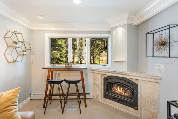 A modern kitchen with a fireplace, a dining area with two stools, and a large window offering a view of trees.