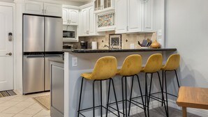 Una cocina con armarios blancos, un refrigerador de acero inoxidable, una cafetera y una zona con taburetes de bar.