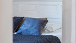 Un lit avec une tête de lit blanche et une literie bleue, une œuvre d’art encadrée au-dessus, et une porte blanche en arrière-plan.