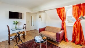 Un salon chaleureux avec un canapé, une table à manger et des chaises, un téléviseur fixé au mur et une fenêtre ornée de rideaux orange.