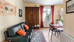 A living room with a black leather sofa, a wooden cabinet, a dining table with chairs, and a window with red curtains.