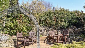 Un espace extérieur aménagé avec des chaises et une table en bois, entouré de verdure et d’un mur en pierre.