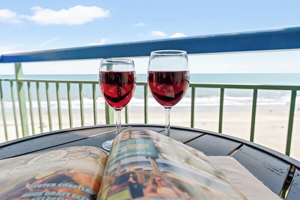 Two glasses of red wine on a table with a magazine, overlooking a beach.