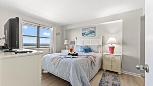 Un dormitorio con cama, mesitas de noche, televisor, ventana con vistas y una puerta.