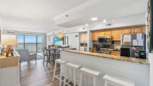 A modern kitchen with granite countertops, wooden cabinets, and stainless steel appliances. There is a dining area with a table and chairs, and a balcony with a view. - Oceans 914 -Oceanfront 2BR- Accommodates 8 (North Myrtle Beach)