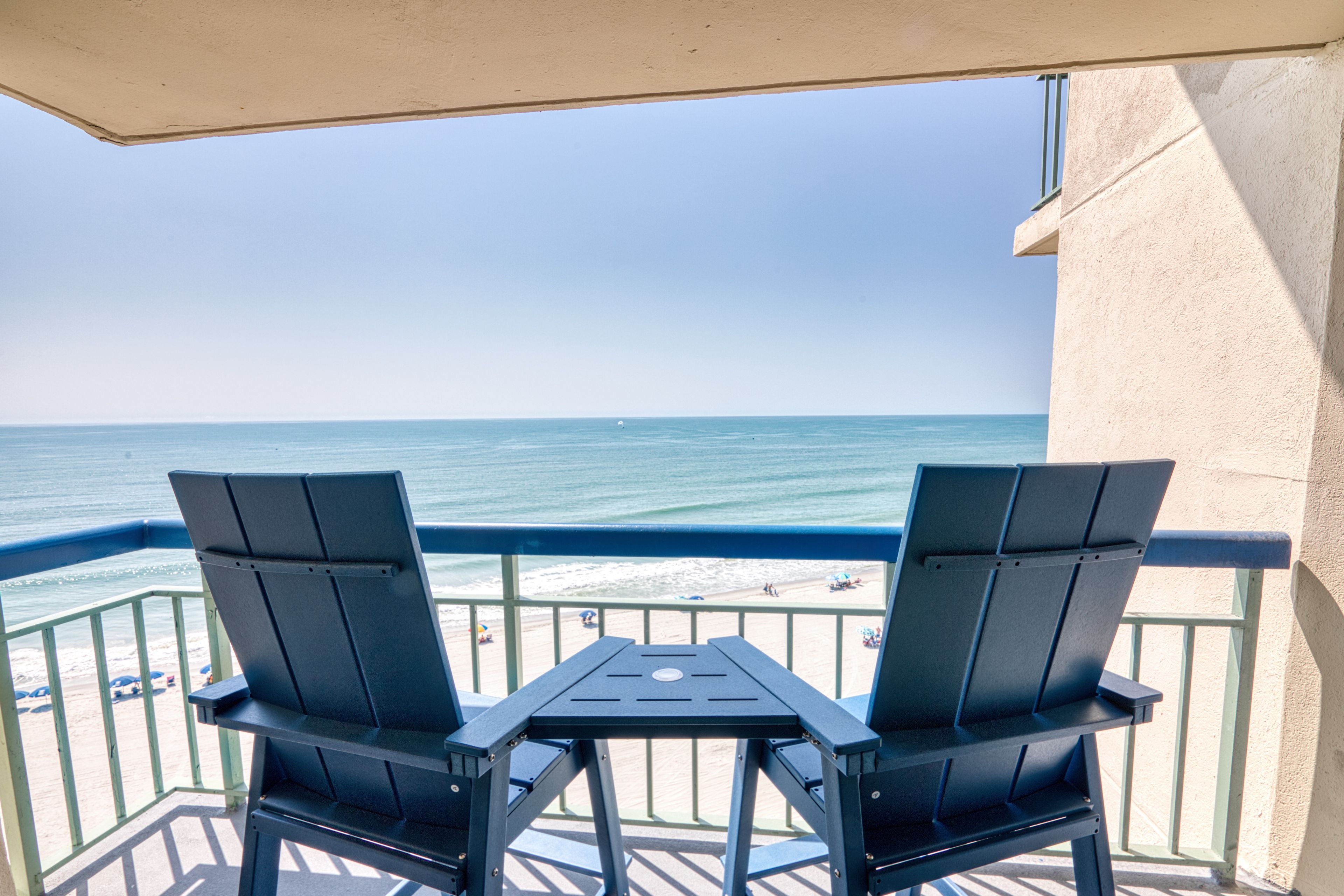 Ein Balkon mit zwei Stühlen und einem Tisch, der auf einen Strand mit dem Meer und einem klaren Himmel blickt.