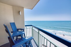 A balcony with two blue chairs and a table, overlooking a beach with people and umbrellas.