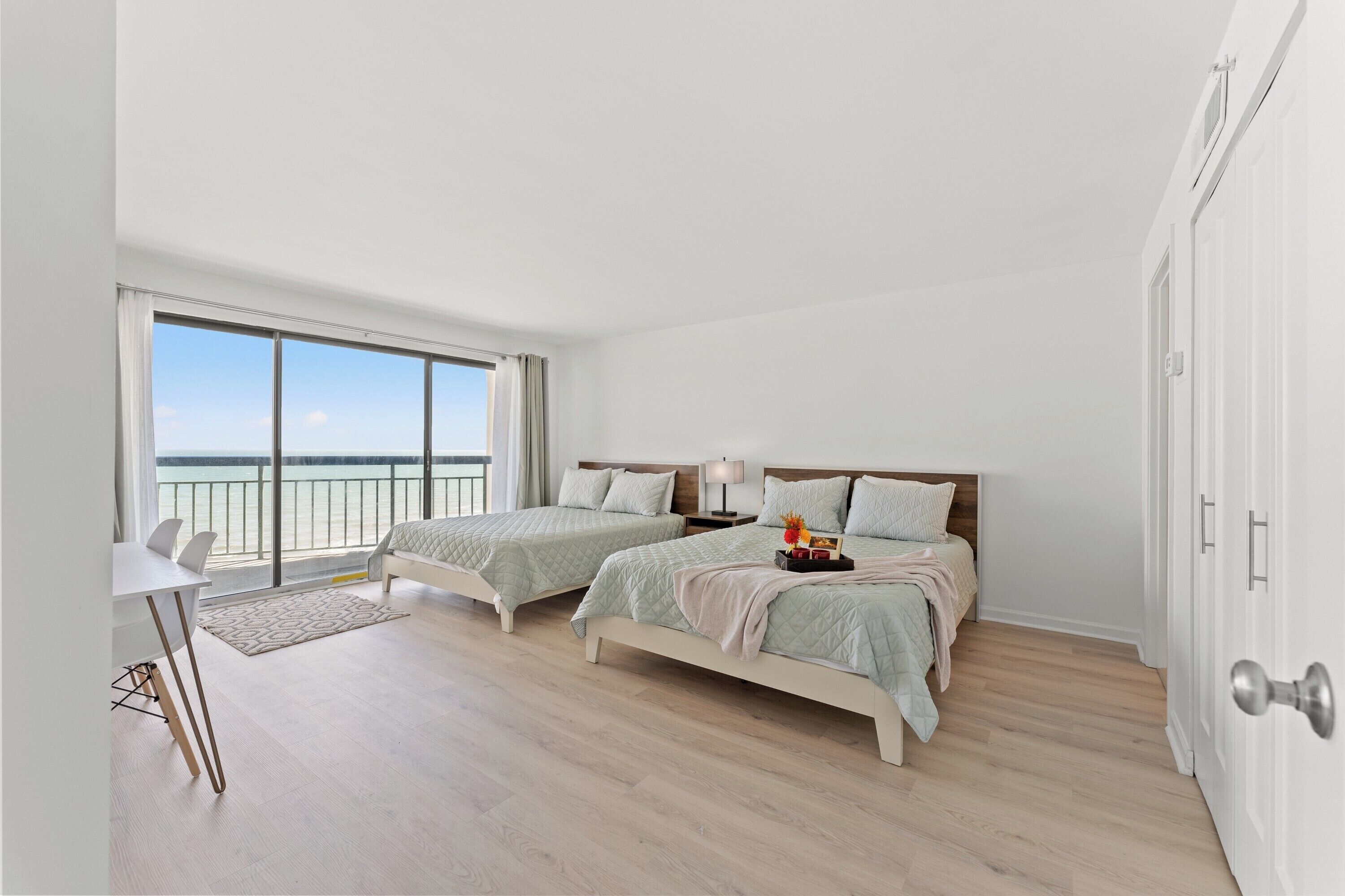 A bedroom with two beds, a desk, and a large window with a view of the sea.