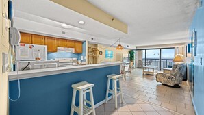 A modern kitchen with a blue accent wall, wooden cabinets, and a bar area with stools. - Oceans 1010 - Ocean Front 1 Bedroom - Pool - Cherry Grove (North Myrtle Beach)