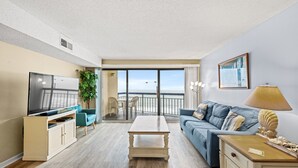 A modern living room with a flat-screen TV, a sofa, a coffee table, and a balcony with a view. - Oceans 1010 - Ocean Front 1 Bedroom - Pool - Cherry Grove (North Myrtle Beach)