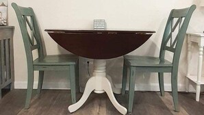 A dining table with a dark top and white base, flanked by two green chairs with a framed picture above. - Steps from Wells Beach, Bellevue by the Sea is perfect for your Maine vacation (Wells)