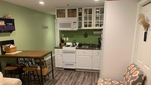 A compact kitchen with white cabinets, a stove, and a microwave. There is a dining table with chairs and a patterned ottoman. - ONE MINUTE WALK to WELLS BEACH, Bellevue by the Sea is your perfect vacation! (Wells)