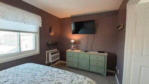 Un dormitorio con cama, cómoda, televisor montado en la pared y ventana con cortina.