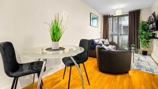 Een moderne woonkamer met een glazen eettafel, zwarte fauteuils, een donkere bank en een groot raam met gordijnen.