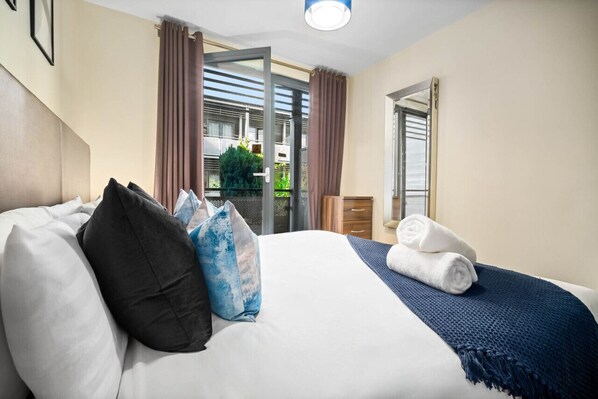 A hotel room with a bed, pillows, a blue blanket, and a view of a balcony with curtains.