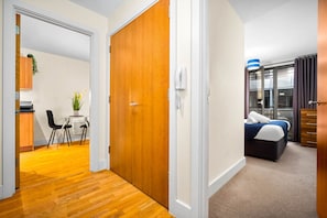A hotel room with a wooden door, a bed with a blue blanket, a desk with a chair, and a small table with a plant.