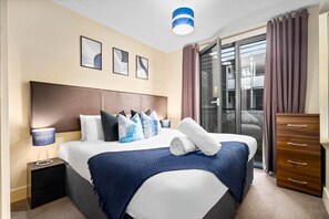 Ein modernes Hotelzimmer mit einem großen Bett, einem hölzernen Kopfteil, einem Nachttisch, einer Kommode und einer Schiebetür zum Balkon.