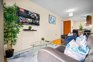 Un salon moderne avec une table basse en verre, un canapé agrémenté de coussins à motifs, un téléviseur diffusant un film et une plante en bambou.