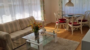 Un salon avec un canapé à rayures, une table basse en verre, une table à manger avec des chaises blanches et des coussins rouges, et un téléviseur posé sur un meuble.