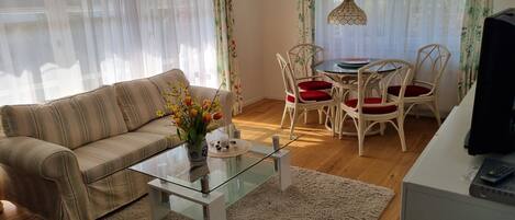 Un salon avec un canapé à rayures, une table basse en verre, une table à manger avec des chaises blanches et des coussins rouges, et un téléviseur posé sur un meuble.