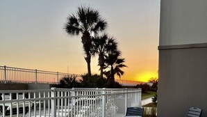 Une terrasse avec une table et des chaises, donnant sur une plage bordée de palmiers et offrant une vue sur le coucher de soleil.