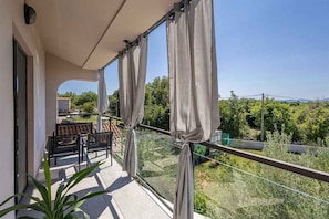 A balcony with a glass railing, a table with chairs, and a view of greenery and buildings.