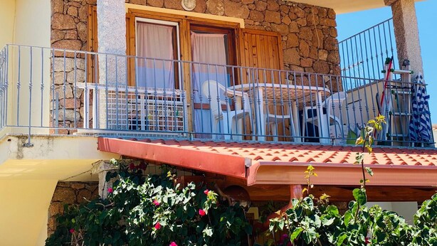 A two-story building with a stone facade, a balcony with a table and chairs, and a red awning. - Villetta 1P NC La Cinta (San Teodoro)