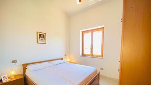 A bedroom with a wooden bed, a nightstand with a lamp, a window with blinds, and a framed picture on the wall. - Villetta 1P NC La Cinta (San Teodoro)