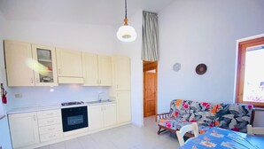 A compact kitchen with white cabinets, a stove, and a sink. There's a wooden door leading to another room, a floral sofa, and a dining table with a blue tablecloth. - Villetta 1P NC La Cinta (San Teodoro)