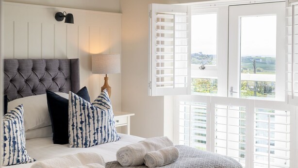 A bedroom with a tufted headboard, a bedside table with a lamp, a bed with rolled towels, and a view through open shutters.
