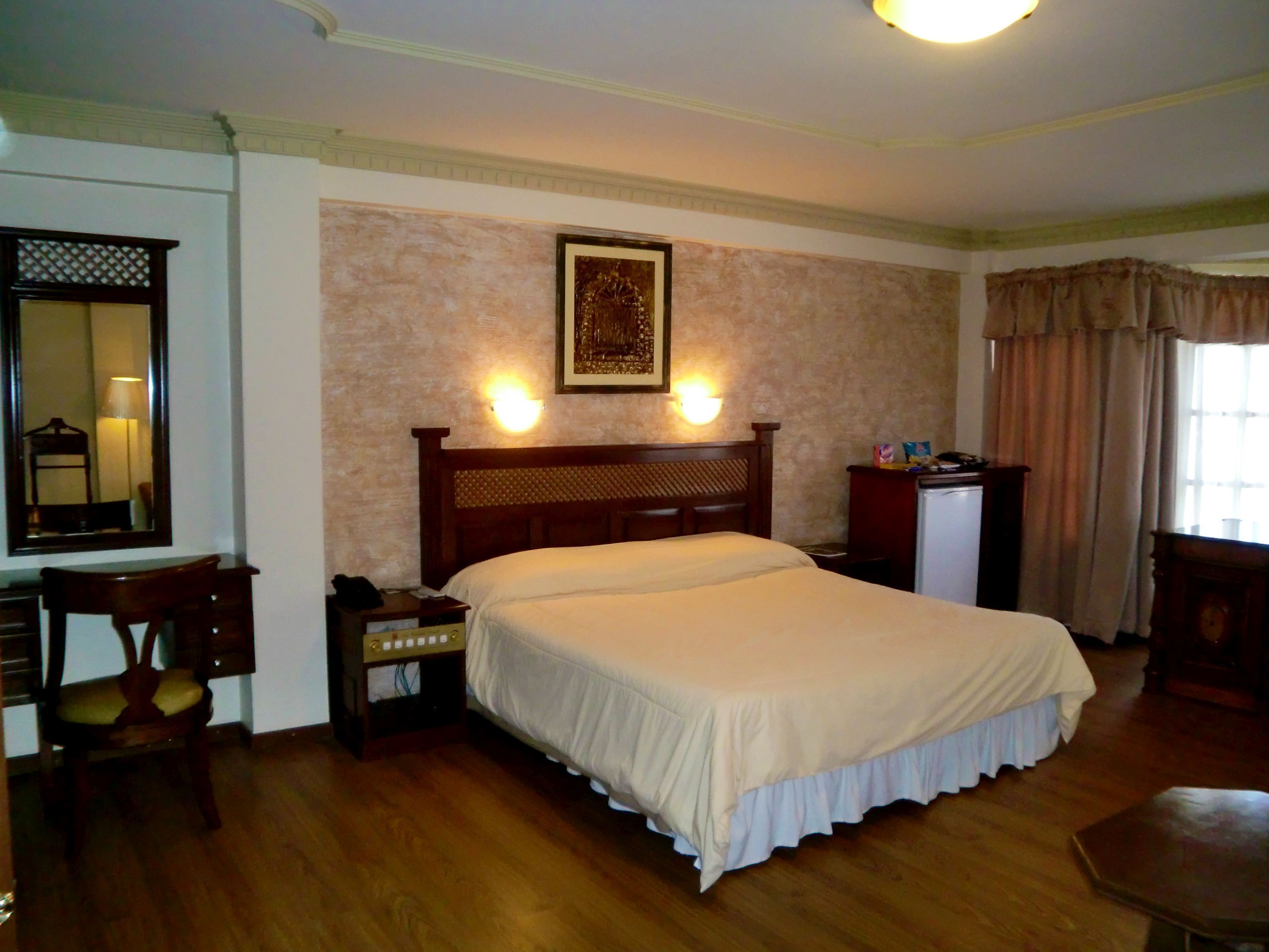 Ein Hotelzimmer mit einem großen Bett, einem Schreibtisch mit Stuhl, einem Schminktisch, einem Spiegel und einer Wandleuchte.