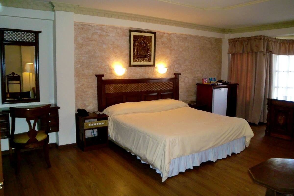 A hotel room with a large bed, a desk with a chair, a dresser, a mirror, and a wall-mounted light fixture.