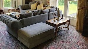 A traditional living room with a large sofa, a matching ottoman, a small table with a candelabra, and a chandelier. There are two windows with curtains, a statue on a side table, and a framed picture on the wall. - Historic Manor House - Sleeps 16 - Pool & Hot Tub (Rochester)