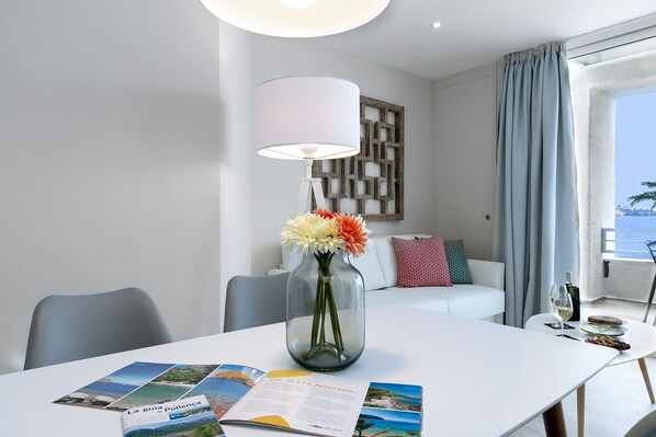 A modern hotel room with a white dining table, a vase with flowers, a magazine, and a view of the sea through a window. - Nice beachfront apartment in Puerto Pollensa (Port de Pollença)