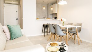 A modern living room with a sofa, a dining table with chairs, and a kitchen area with a refrigerator and cabinets. - Nice beachfront apartment in Puerto Pollensa (Port de Pollença)