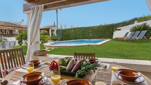 Un espacio de comedor al aire libre con vista a la piscina, muebles de madera y una variedad de frutas y verduras sobre la mesa.