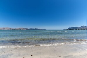 Ein Strand mit klarem Wasser, sandigem Ufer und Bergen in der Ferne.