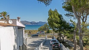 Una strada costiera con auto parcheggiate, un edificio bianco con antenna parabolica e una vista aperta sul mare e sulle montagne sullo sfondo.