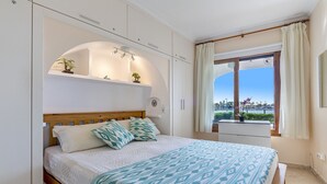 A bedroom with a bed, a window with curtains, a view of the outdoors, and built-in shelves with plants. - 1st line apartment in Alcudia Beach CAR (Alcúdia)