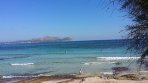 Uma praia com águas cristalinas, montanhas ao longe e algumas pessoas nadando.