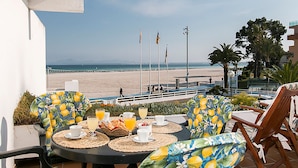 A balcony with a table set for breakfast, overlooking a beach with flags, palm trees, and a clear sky. - Seafront apartment in Alcudia beach (Port d´Alcudia)