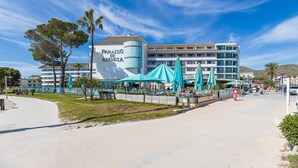 A hotel with a prominent sign, outdoor seating with umbrellas, and a clear sky. - Seafront apartment in Alcudia beach (Port d´Alcudia)