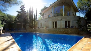 Una casa de dos pisos con piscina, paredes de piedra y un balcón con sillas.