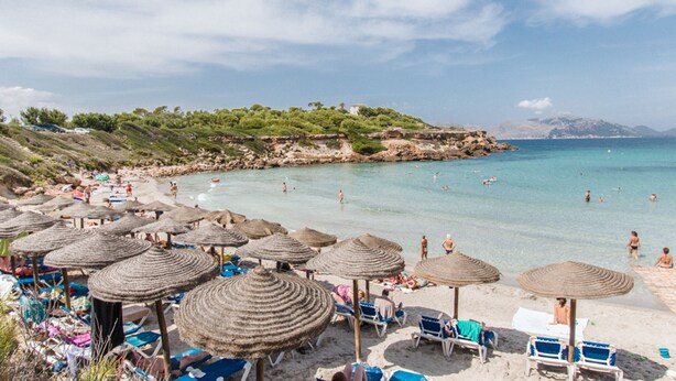 Una playa con sombrillas de paja, sillas de tomar el sol y gente nadando.
